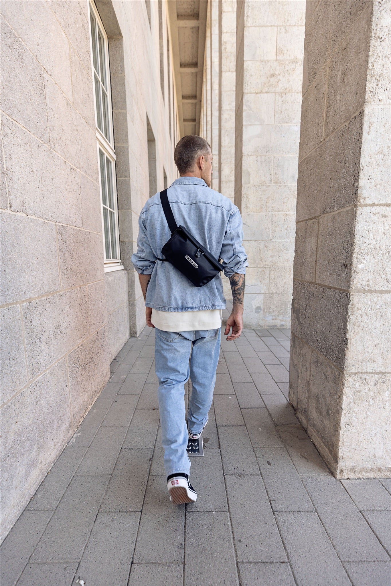 Vista trasera de hombre con bandolera negra ORTLIEB Velo-Sling Flex con cierre enrollable caminando por columnata.